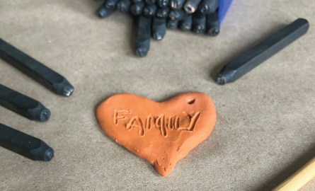 Two pieces of terracotta airdry clay, one in the shape of a heart and one in the shape of a rectangle, is surrounded by letter stamps and a pointed wooden pick. On the heart-shaped piece of clay, the word "Family" is etched into it. On the rectangle piece of clay, the words "love you" are stamped into it.